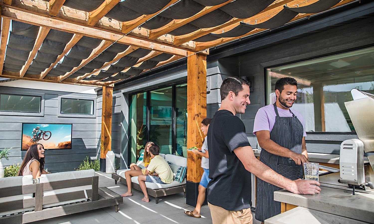 A luxurious outdoor entertainment space featuring a large wall-mounted TV, sleek modern design, and built-in fire pits surrounded by lush greenery.