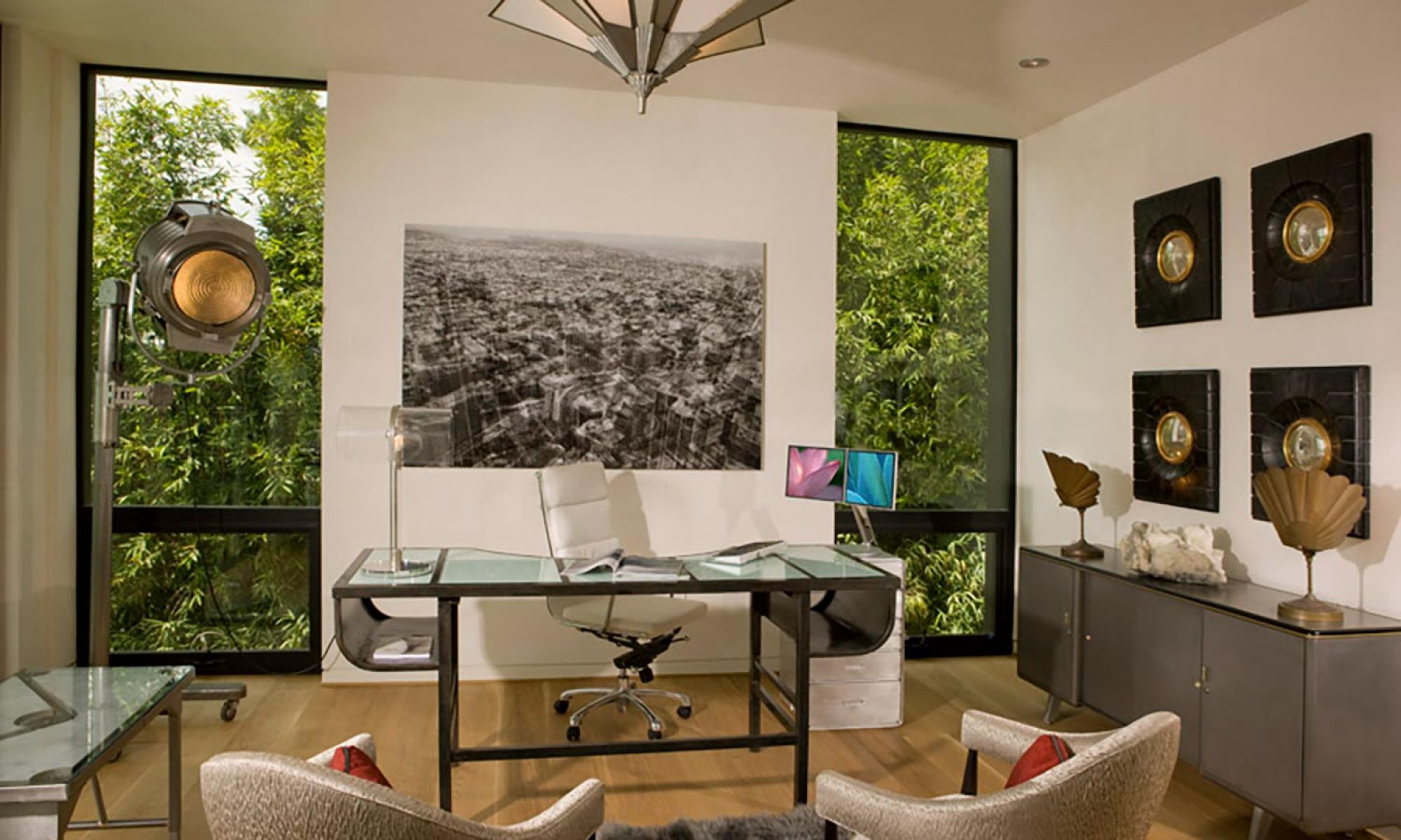 A stylish home office with a glass desk, a large black-and-white cityscape photo, and unique wall art. A vintage spotlight and lush greenery outside the windows add character.