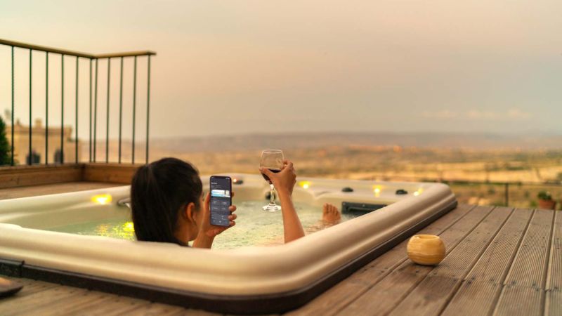 A woman relaxing in an outdoor hot tub at sunset, holding a glass of wine in one hand and a smartphone displaying a smart home control app in the other, overlooking a scenic landscape.
