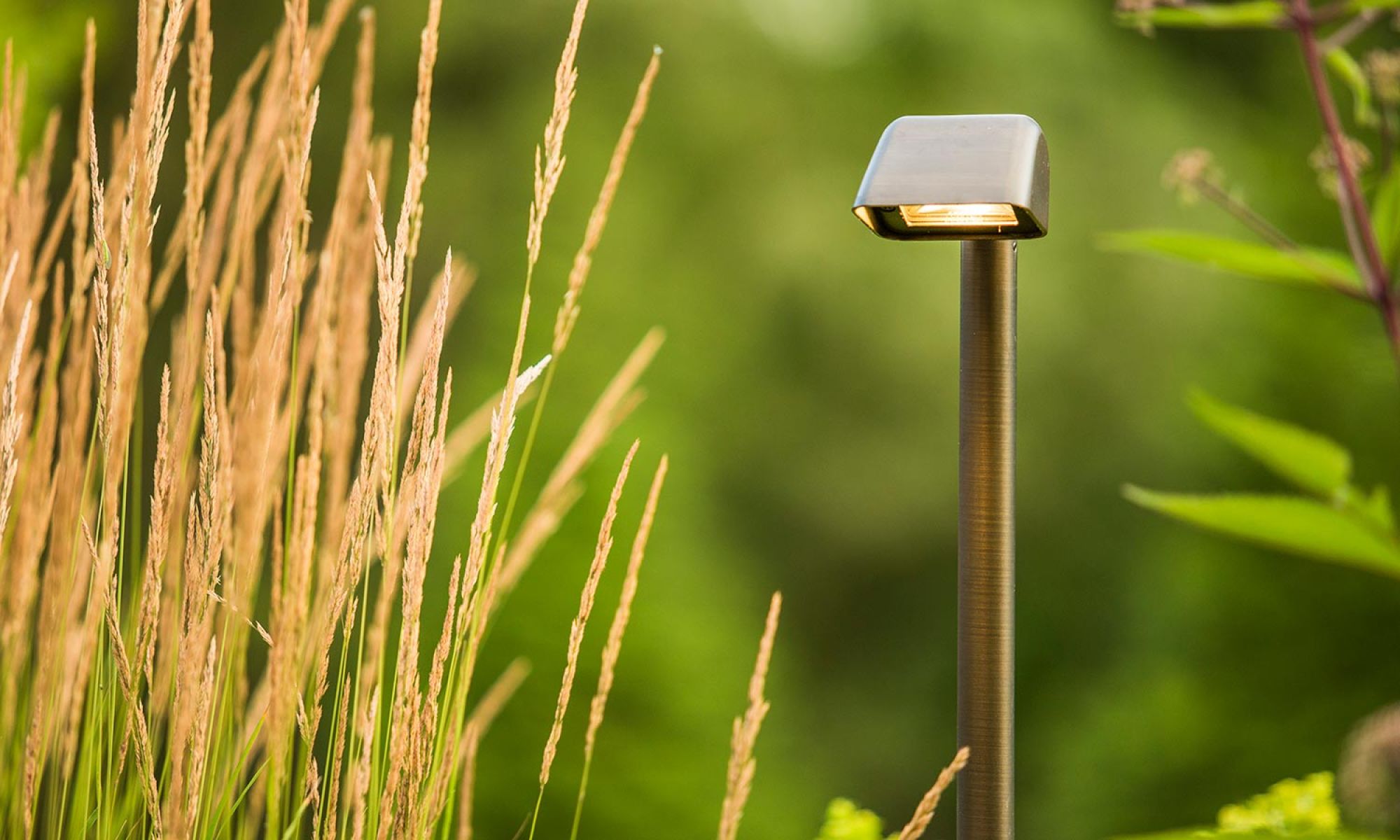 A modern landscape lighting fixture with a sleek, angular design, standing tall among golden ornamental grasses and lush greenery.