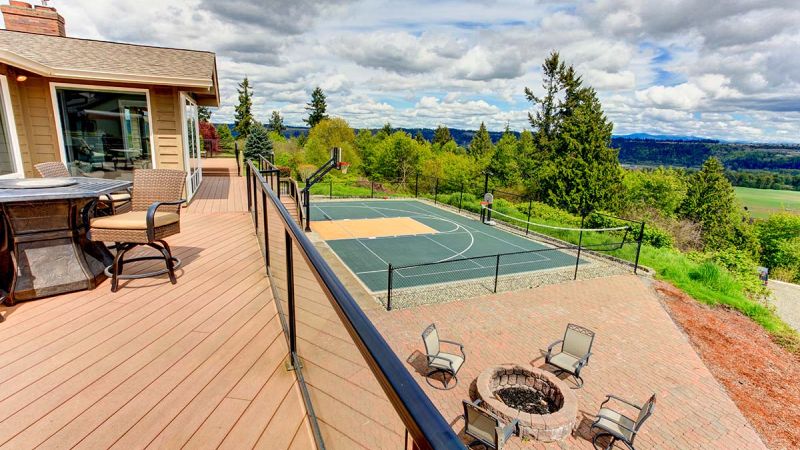 A scenic backyard with a spacious wooden deck, a fire pit surrounded by chairs, and a full-size basketball court, all overlooking a lush green valley.