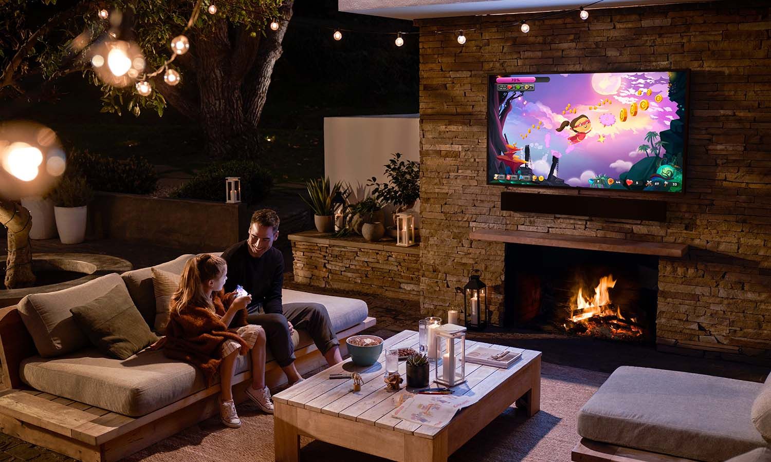 A father and daughter sitting on a cozy outdoor couch at night, enjoying quality time near a fireplace with string lights above, while a TV on the stone wall displays a colorful video game.