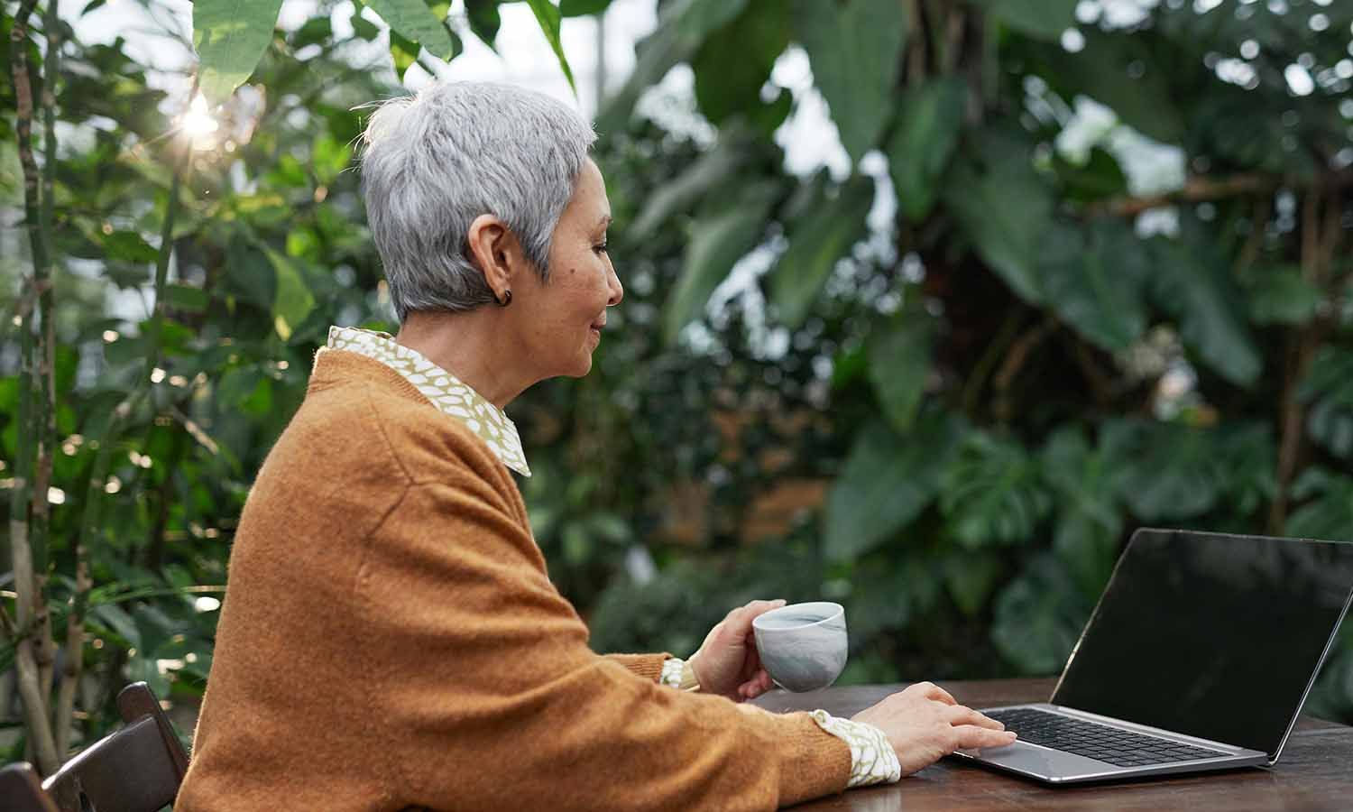 A woman with short gray hair sits at a wooden table in a lush green outdoor setting, enjoying a cup of coffee while working on her laptop.